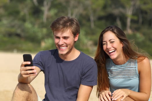 Funny man and woman laughing watching the smart phone on the beach on summer holidays