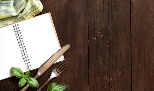 Blank cooking recipe book with fork, knife, basil and napkin on a wooden table