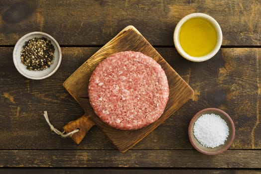 Burger patty from ground beef on wooden board with olive oil, salt and pepper