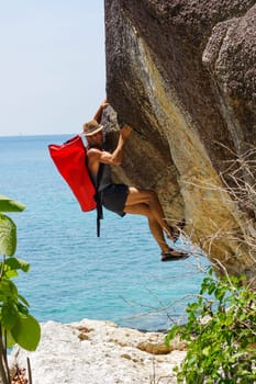 a strong and happy man climbing on high rock over the sea with a hut and a red seabag.
