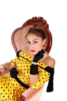 A gorgeous young woman sitting in a pink armchair in a yellow dress and.wearing black cloves and jewellery, isolated for white background..