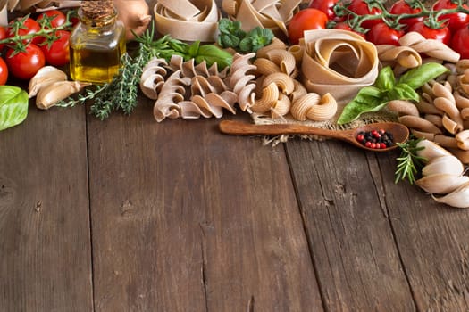 Whole wheat pasta, vegetables, herbs and olive oil on wooden background