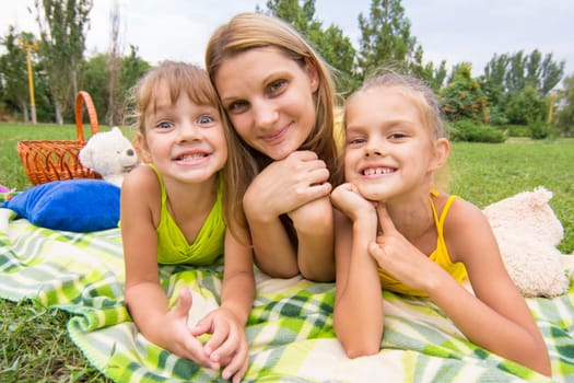 Mother and two girls lying on the grass on a picnic and fun look into the frame