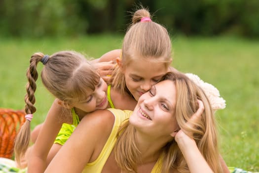 Two daughter kissing and hugging her mother lying on the grass on a picnic