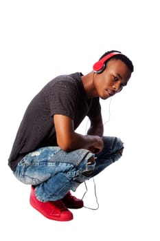 Handsome African teenager listening to music wearing red headphones while squatting, on white.