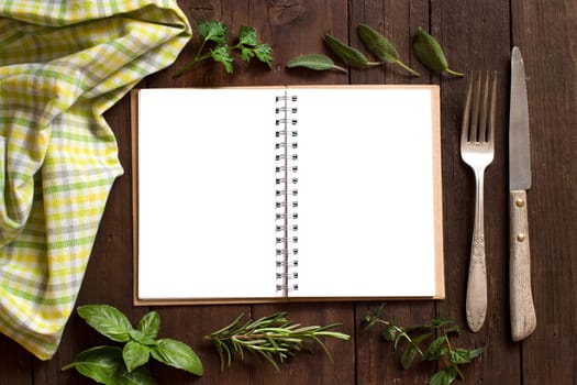 Blank cooking recipe book with fork, knife, herbs and napkin on a wooden table