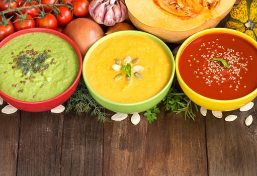 Three fresh soups in colorful bowls and vegetables on a wooden table