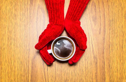 Woman hands in mittens holding a cup of coffee on wooden background