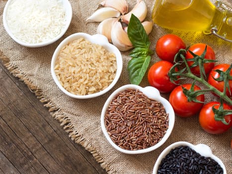 Variety of rice in bowls, garlic, basil, olive oil and tomatoes on wooden table