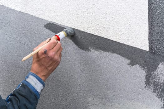 Close-up arm of a painter with paint roll in hand paints a house wall with gray paint.