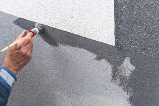 Close-up arm of a painter with paint roll in hand paints a house wall with gray paint.