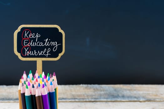 Wooden mini blackboard label with message Key Keep Educating Yourself and colouring pencils over chalkboard background, copy space available, education concept