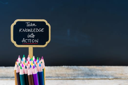 Wooden mini blackboard label with message Turn Knowledge Into Action and colouring pencils over chalkboard background, copy space available, education concept
