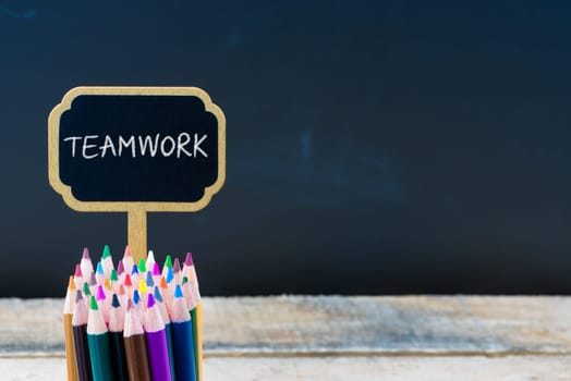 Wooden mini blackboard label with message TEAMWORK and colouring pencils over chalkboard background, copy space available, education concept