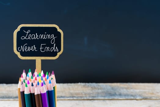 Wooden mini blackboard label with message LEARNING NEVER ENDS and colouring pencils over chalkboard background, copy space available, education concept