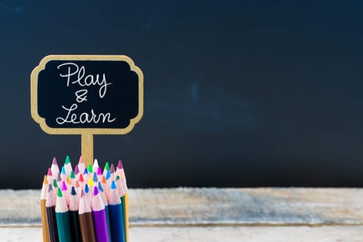 Wooden mini blackboard label with message Play and Learn and colouring pencils over chalkboard background, copy space available, education concept