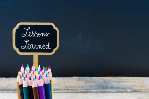 Wooden mini blackboard label with message Lessons Learned and colouring pencils over chalkboard background, copy space available, education concept