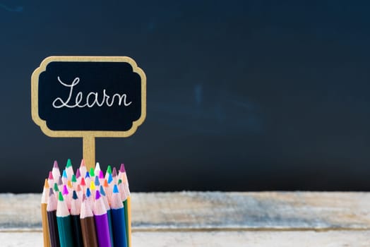 Wooden mini blackboard label with message LEARN and colouring pencils over chalkboard background, copy space available, education concept