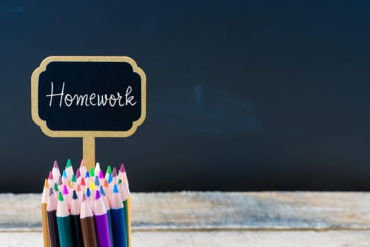 Wooden mini blackboard label with message HOMEWORK and colouring pencils over chalkboard background, copy space available, education concept