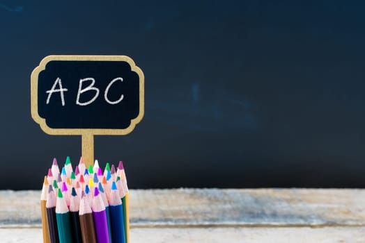 Wooden mini blackboard label with message ABC and colouring pencils over chalkboard background, copy space available, education concept