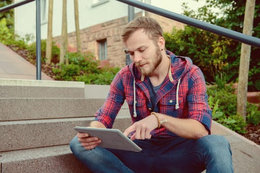 young man in checkered shirt sitting on stairs and using tablet PC