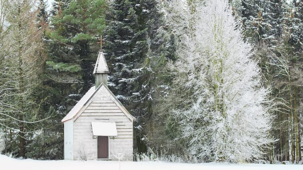 Peaceful winter view of little snow-capped rural wooden church on snowfield glade in frozen forest