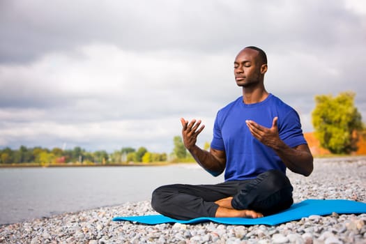 young black man wearing athletic wear sitting on the beach exercising yoga