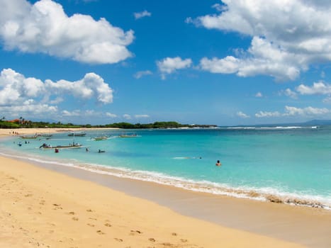 Beautiful white sand beach surrounded by tropical palm trees on the island of Bali in Nusa Dua area. Blue sky with white clouds, white sand and boats