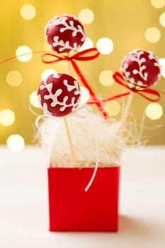 Red cake pops garnished with white icing sticking in red gift box. Very shallow depth of field with Christmas lights in the background.
