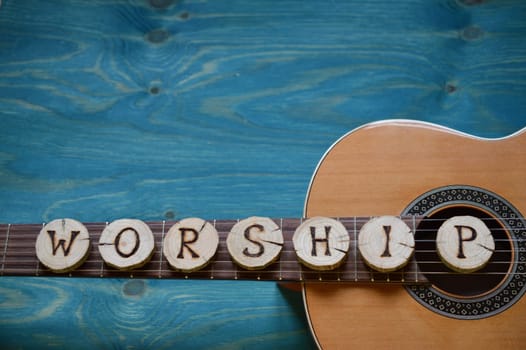 Guitar on blue-green wood with individual wooden discs on it,which form the word: WORSHIP