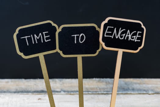 Business message TIME TO ENGAGE written with chalk on wooden mini blackboard labels, defocused chalkboard and wood table in background