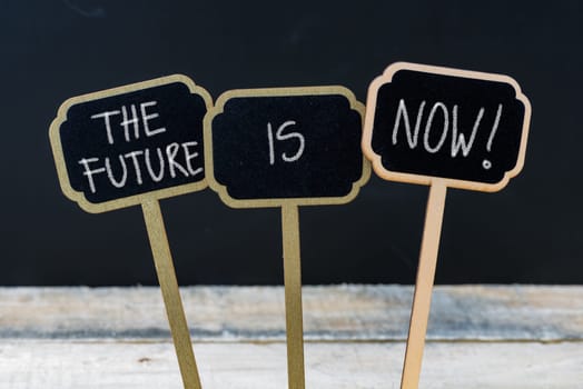 Business message THE FUTURE IS NOW written with chalk on wooden mini blackboard labels, defocused chalkboard and wood table in background