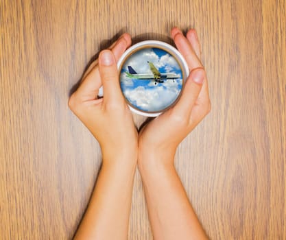 Woman holding cup with dream about trip or vacation by airplane against wooden table. Top view. Conceptual photo.