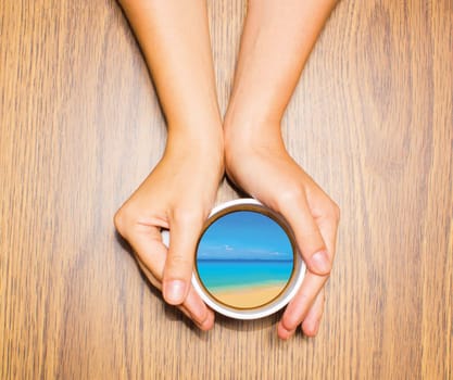Woman holding cup with dream about tropical vacation against wooden table. Top view. Conceptual photo. Collage