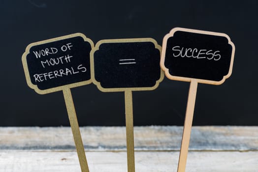 Business message WORD OF MOUTH REFERRALS EQUALS SUCCESS written with chalk on wooden mini blackboard labels, defocused chalkboard and wood table in background