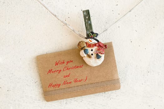 Snowman clothespins hanging on clothesline or rope and holding Christmas greeting card on plaster background