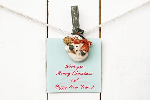 Christmas snowman clothespins hanging on clothesline or rope and holding light blue greeting note paper card on wooden background