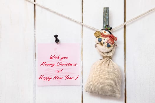 Christmas snowman clothespin hanging on clothesline or rope and holding sack and pink Christmas greeting note paper with push black pin on wood background