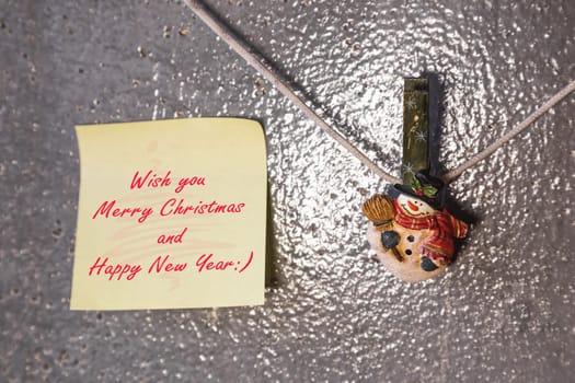 Christmas snowman clothespins hanging on clothesline or rope and yellow greeting note paper card on plaster background