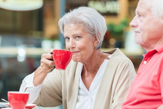 Two smiled seniors sit in a café and drink from a red cup