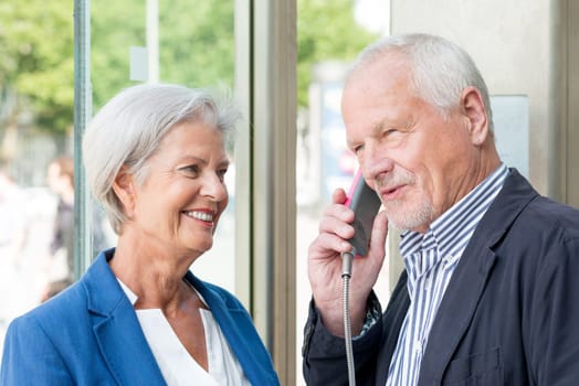 Two seniors stand at a public telephone booth and make phone calls