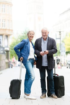 Two seniors with suitcases on a sidewalk and cell phones