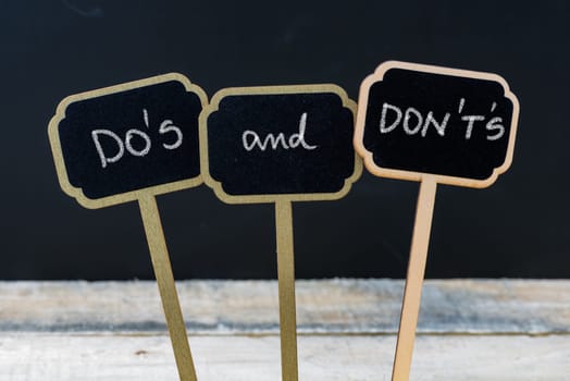 Business message DO'S and DON'T'S written with chalk on wooden mini blackboard labels, defocused chalkboard and wood table in background