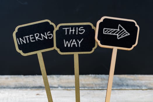 Business message INTERNS THIS WAY written with chalk on wooden mini blackboard labels, defocused chalkboard and wood table in background