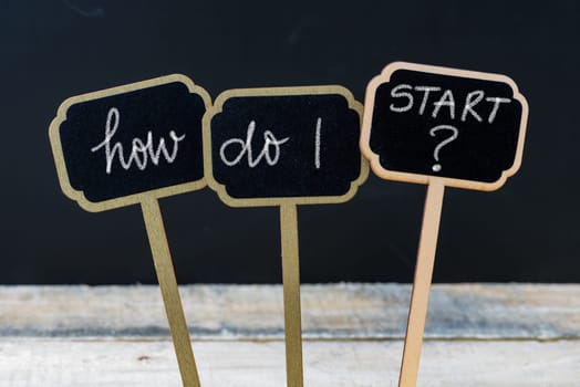 Business message HOW DO I START ? written with chalk on wooden mini blackboard labels, defocused chalkboard and wood table in background