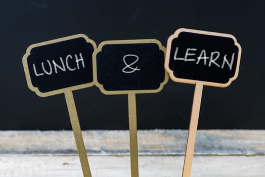 Business message LUNCH and LEARN written with chalk on wooden mini blackboard labels, defocused chalkboard and wood table in background