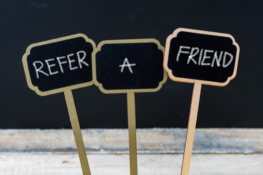 Business message REFER A FRIEND written with chalk on wooden mini blackboard labels, defocused chalkboard and wood table in background