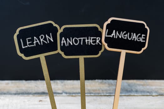 Business message LEARN ANOTHER LANGUAGE written with chalk on wooden mini blackboard labels, defocused chalkboard and wood table in background
