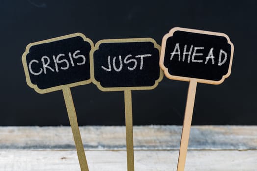 Business message CRISIS JUST AHEAD written with chalk on wooden mini blackboard labels, defocused chalkboard and wood table in background