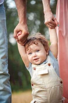 Young beautiful father, mother and little toddler son against green trees. Happy family with her baby boy on a summer sunny day. Family walking on the meadow.